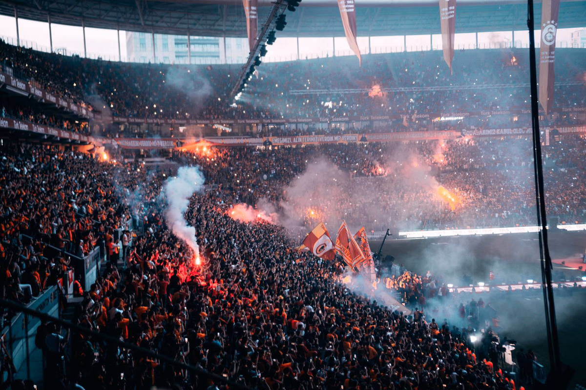 A packed night stadium as fans light flares and wave flags through the smoke at a major match