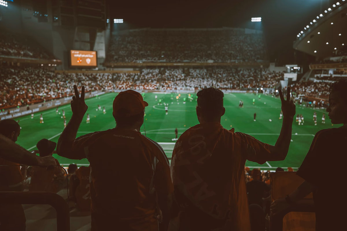 แฟนบอลยืนชมเกมจากอัฒจันทร์ในสนามฟุตบอลยามค่ำคืนที่เปิดไฟสว่าง