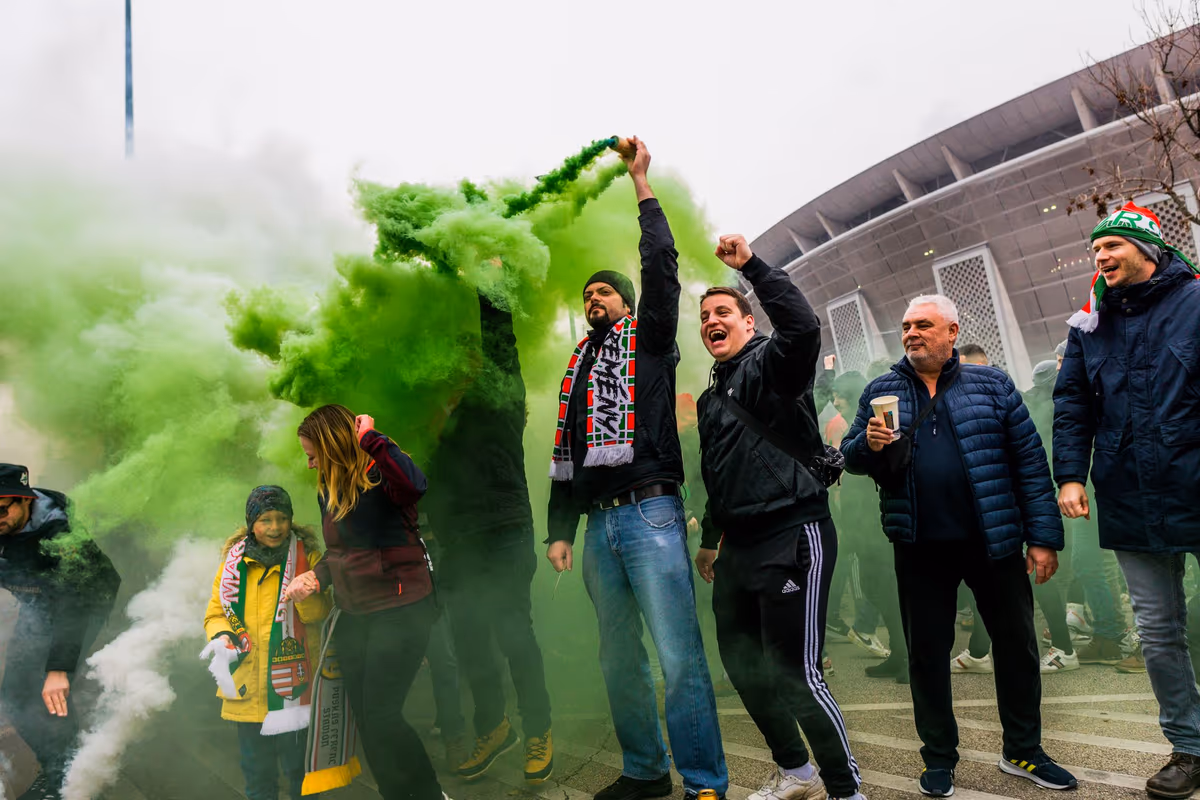 Football fans lighting green smoke and raising their arms in celebration outside a stadium