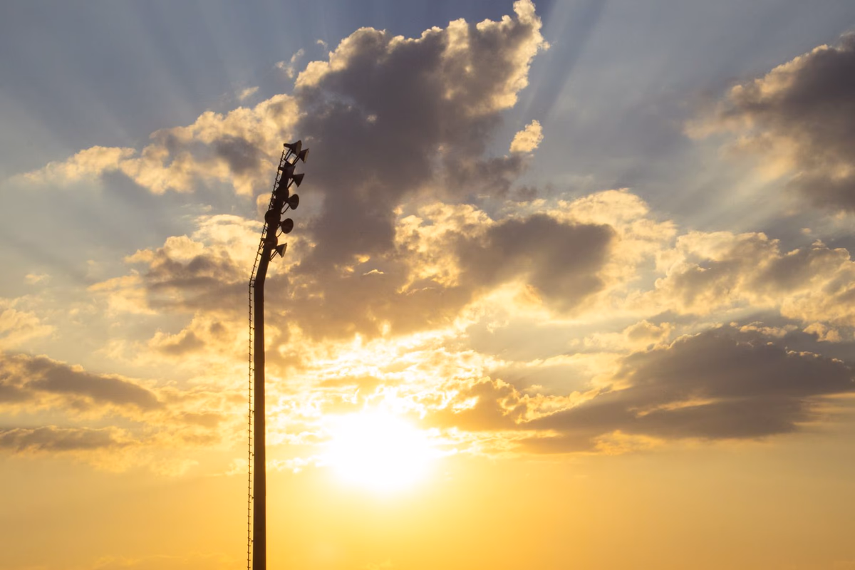 A stadium floodlight tower silhouetted against a burning golden sunset, light breaking through the clouds before an evening match