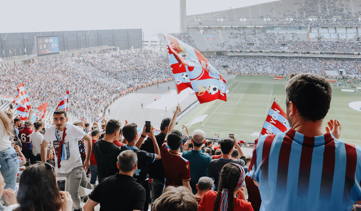 Football supporters fill a packed stadium, waving flags and roaring at a major knockout match