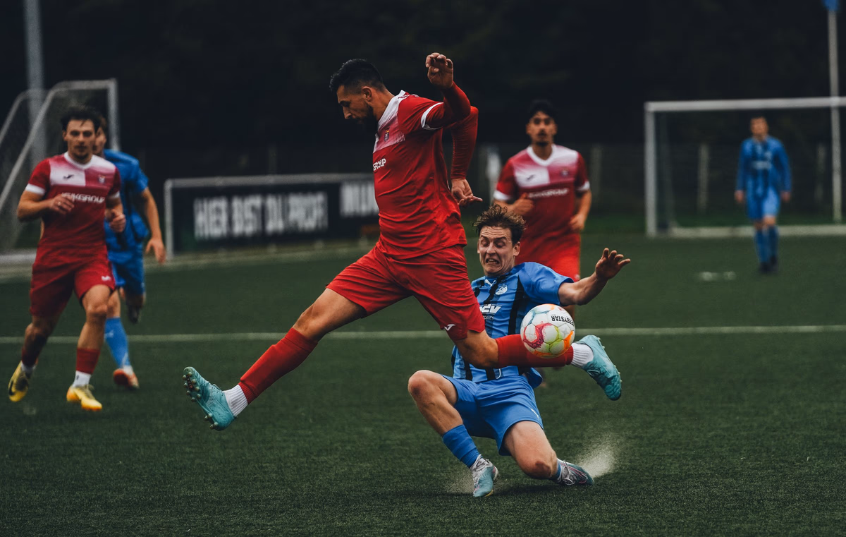 Two footballers, one in red and one in blue, fly into a fierce challenge for the ball in midfield