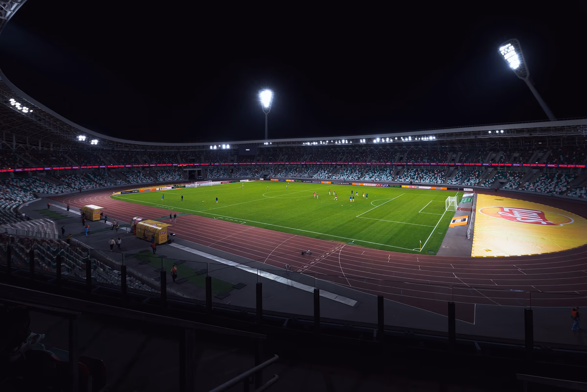 A floodlit football stadium at night with players on the green pitch