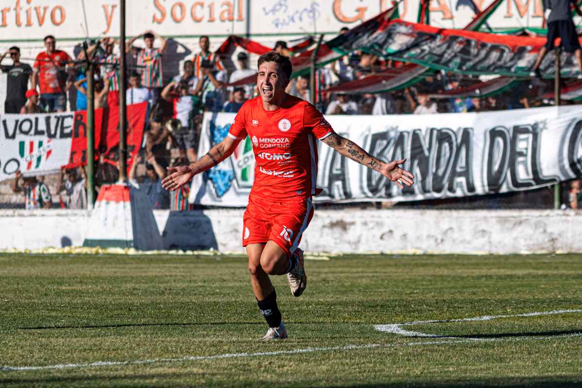 A footballer in a red kit runs with arms outstretched celebrating a goal in front of a packed terrace