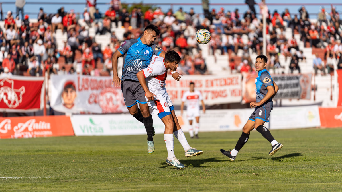 Two players leaping to contest an aerial header inside the penalty area with a crowd behind