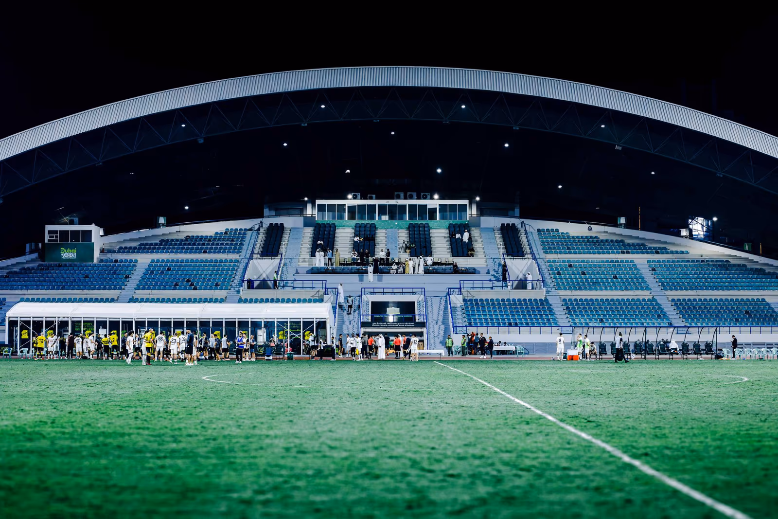 A modern football stadium under floodlights at night with players warming up by the touchline